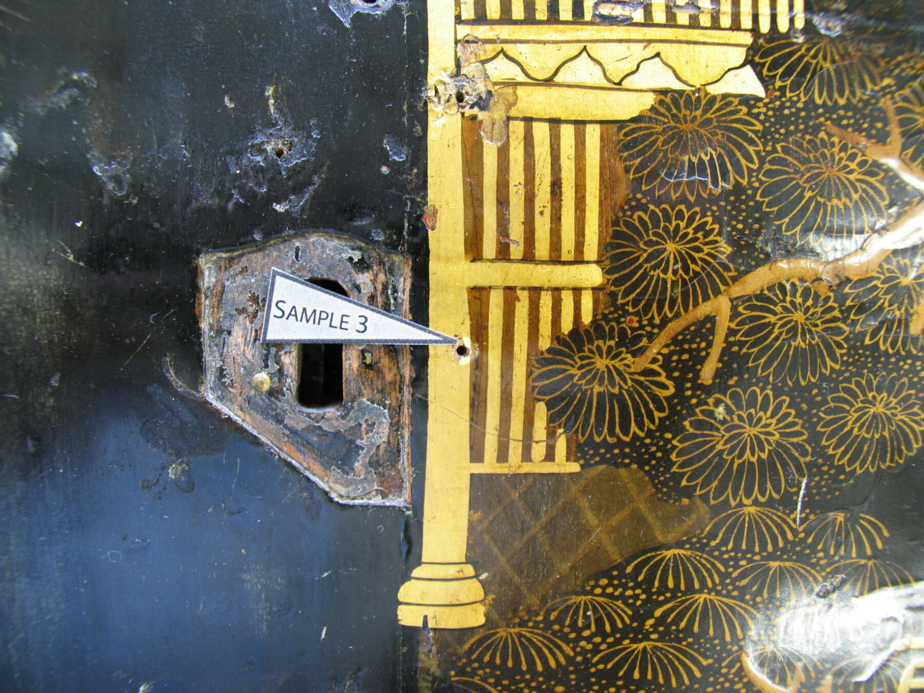 detail of one of the lacquer scenes, with a white triangle with black text reading “sample 3” pointing to a hole in one of the pagoda columns where a sample was taken