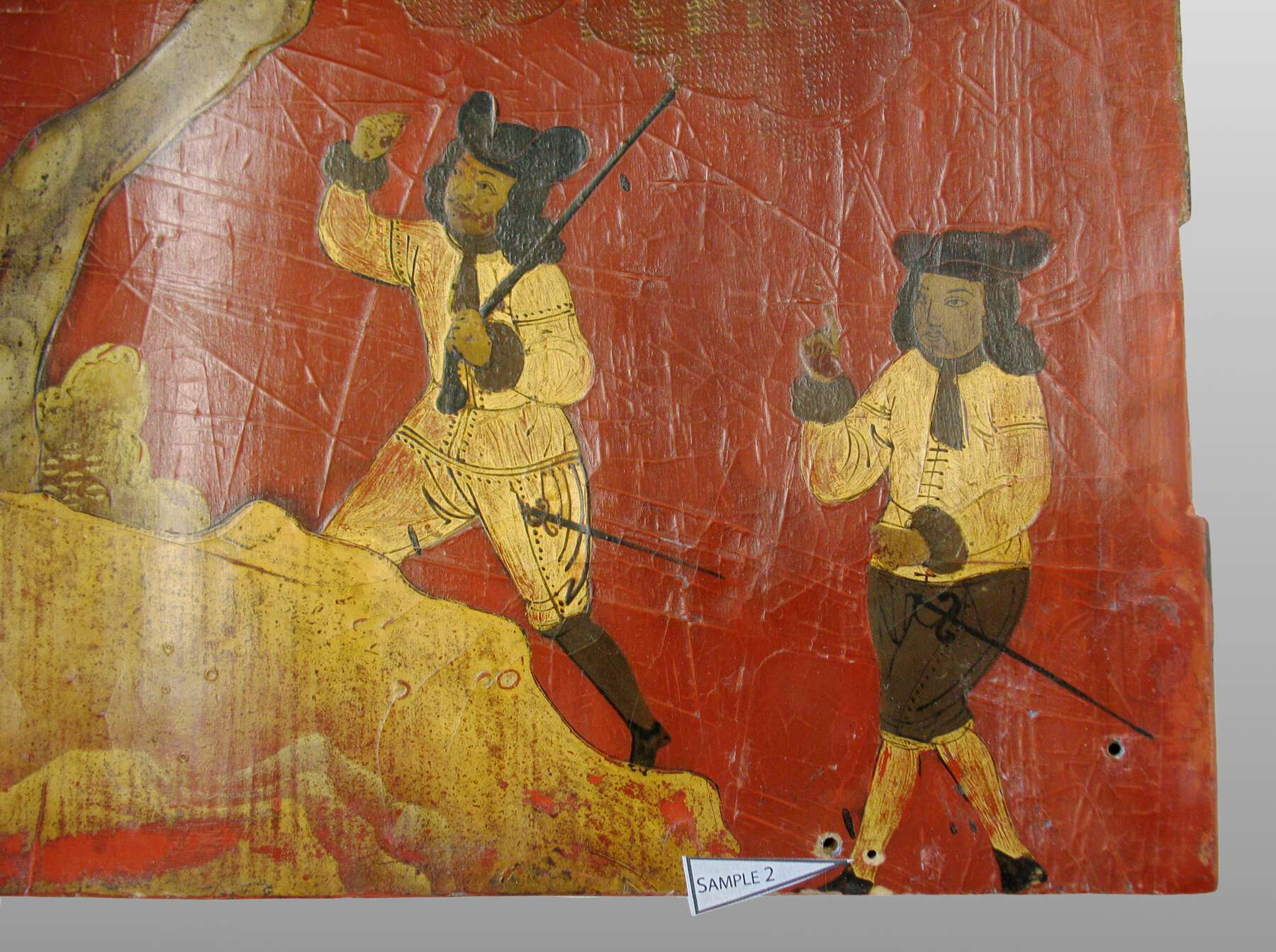 detail of one of the lacquer scenes, with a white triangle with “Sample 2” printed on it pointing to a hole in the ankle of one of the figures