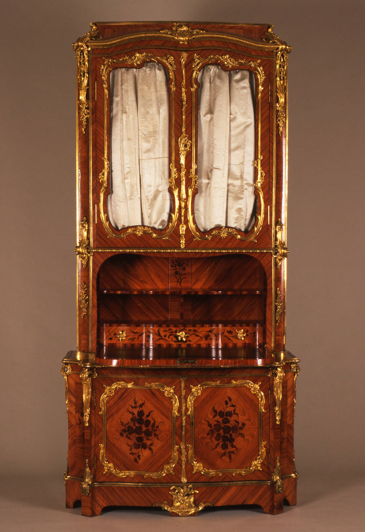 a tall secretary desk with two sets of doors, one under the desktop of shelves and drawers, and the other above, constructed with three different types of wood and decorated with gilt bronze mounts, white marble accents, and two white satin curtains installed inside the upper set of cabinet doors